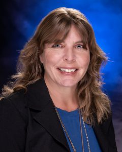 Woman with long wavy brown hair and curtain bangs wears a navy blue shirt under a black blazer